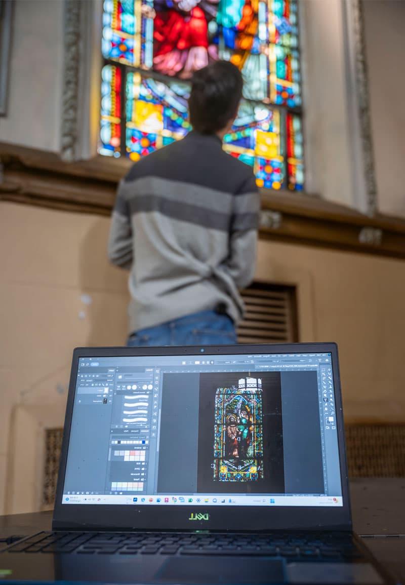 A laptop with an image of a stained glass window is in the foregound, with a male student standing at a stained glass window in the background.