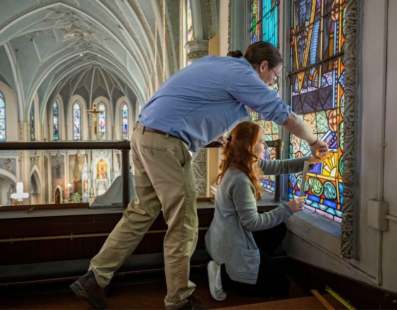 一名女学生跪在彩色玻璃窗底部测量尺寸，而一名男教授站在她上方, helping.